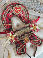 A red and black tartan plaid costume accessory set featuring a large headpiece, a long sash, and two large bows, decorated with gold lace trim, cream ribbons, and central gold and red heart medallions, displayed on a tufted cream upholstered chair.